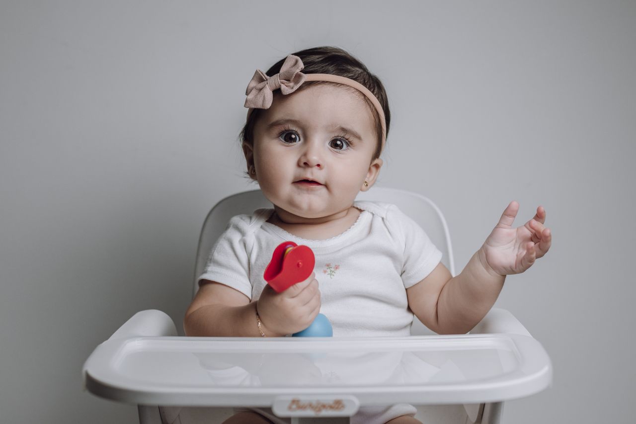 Home Cute baby girl with bow headband sitting in a high chair, playing with a toy.